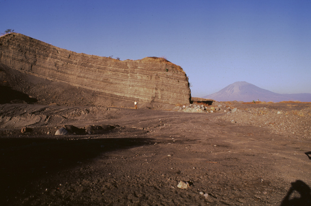 Bedded pyroclastic-surge deposits are exposed in a quarry at Laguna de Aramuaca, SE of the city of San Miguel.  Conical San Miguel volcano rises 18 km to the west in the right background.  Note the person in the center for scale. Photo by Giuseppina Kysar, 1999 (Smithsonian Institution).