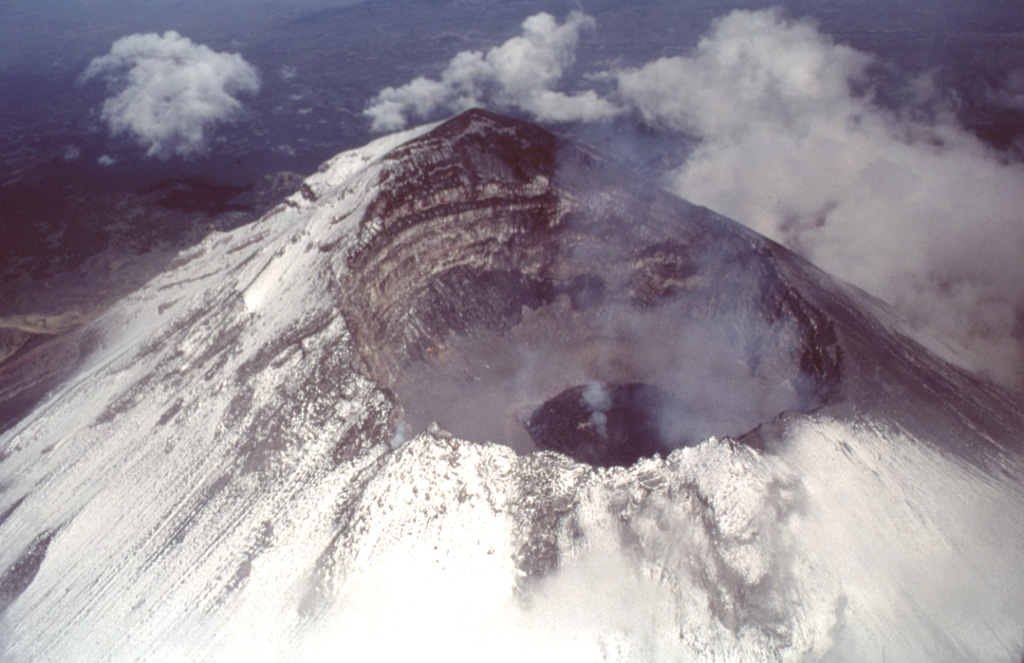 An aerial view of Popocatépetl from the east on 4 July 1997 shows a dark gray lava dome with a strong fumarole emanating from its center. This dome was extruded after an explosive event on 30 June 1997 produced an eruptive column that deposited a thin ash layer on Mexico City. The far crater wall reveals a stratigraphic profile of lava flows and fragmental material forming the upper part of the cone. Photo by José Macías, 1997 (Universidad Nacional Autónoma de México).