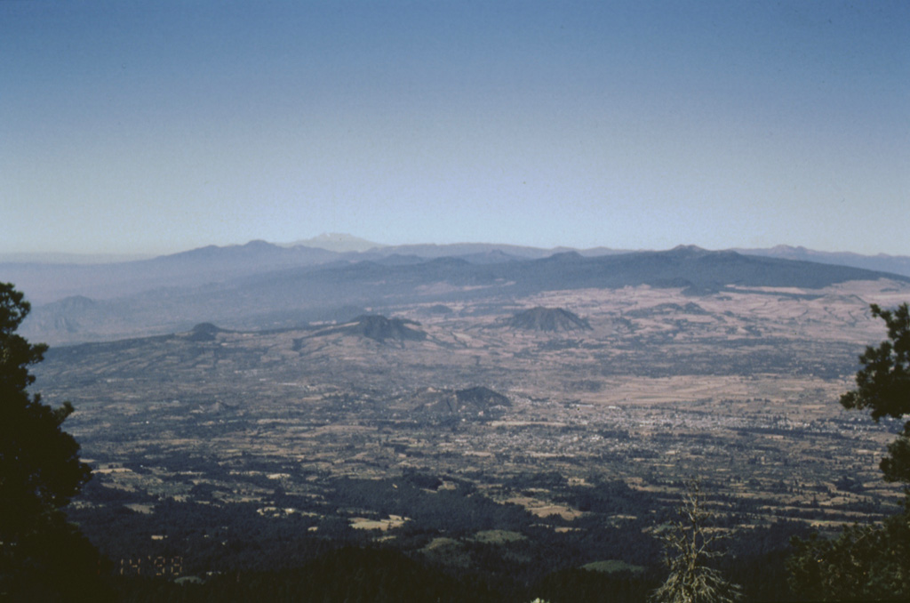 A wide-angle view of the Chichinautzin volcanic field from the flanks of Popocatépetl shows some of the abundant pyroclastic cones and low shield volcanoes that form the mostly monogenetic field. The massive Chichinautzin volcanic field covers more than 1,000 km2 and stretches 90 km in an E-W direction from the eastern base of Nevado de Toluca volcano (the light-colored peak on the far horizon) to the western flanks of Popocatépetl and Iztaccíhuatl. Photo by José Macías, 1998 (Universidad Nacional Autónoma de México).