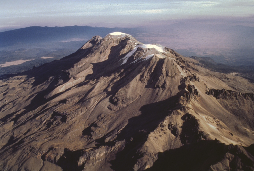 Iztaccíhuatl is a massive 450 km3 stratovolcano SE of Mexico City, seen here from the SW. The summit is composed of several overlapping edifices, including the northernmost peak, La Cabeza (left), the snow-capped high point El Pecho, and Las Rodillas (below the lower Ayoloco glacier near the center). Most activity ceased during the Pleistocene, and the volcano has been extensively glaciated, as seen by the sharp-crested glacial moraines below the Ayoloco glacier. Photo by José Macías, 1995 (Universidad Nacional Autónoma de México).