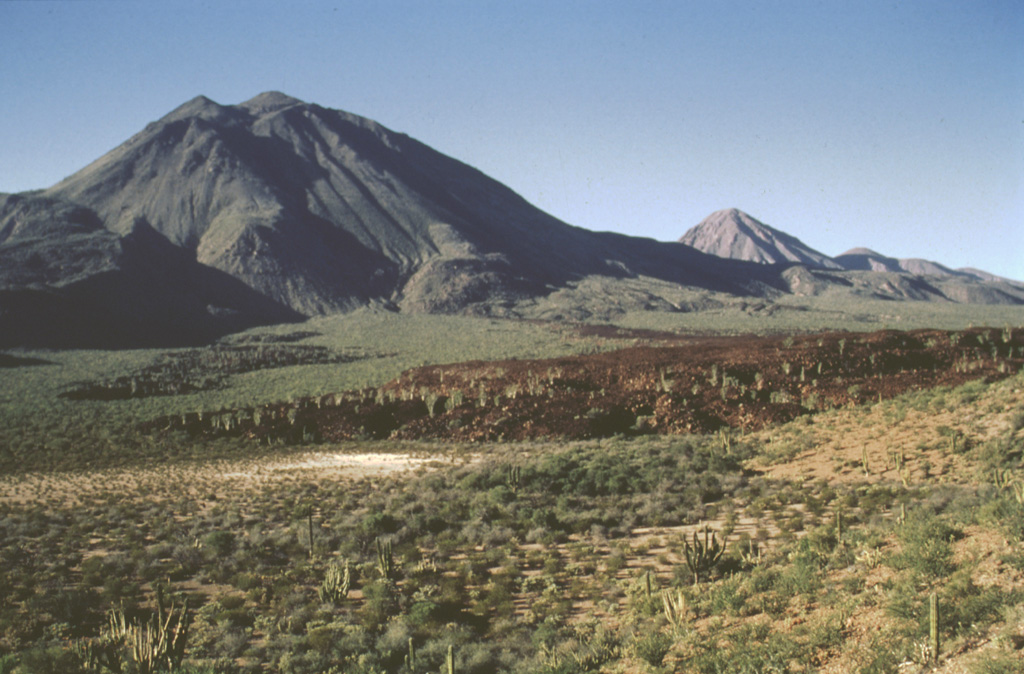 The Tres Vírgenes volcanic complex consists of three stratovolcanoes constructed along a NE-SW line, seen here from the SE. La Vírgen (the youngest and highest when this 1995 photo was taken) is seen to the he left, with the El Azufre peak to the right and the older El Viejo forming the lower peak to the far right. The sparsely vegetated dark lava flow in the foreground is one of the youngest from La Virgen. A major Plinian eruption from a SW-flank vent was followed the by extrusion of a thick lava flow. Photo by José Macías, 1995 (Universidad Nacional Autónoma de México).