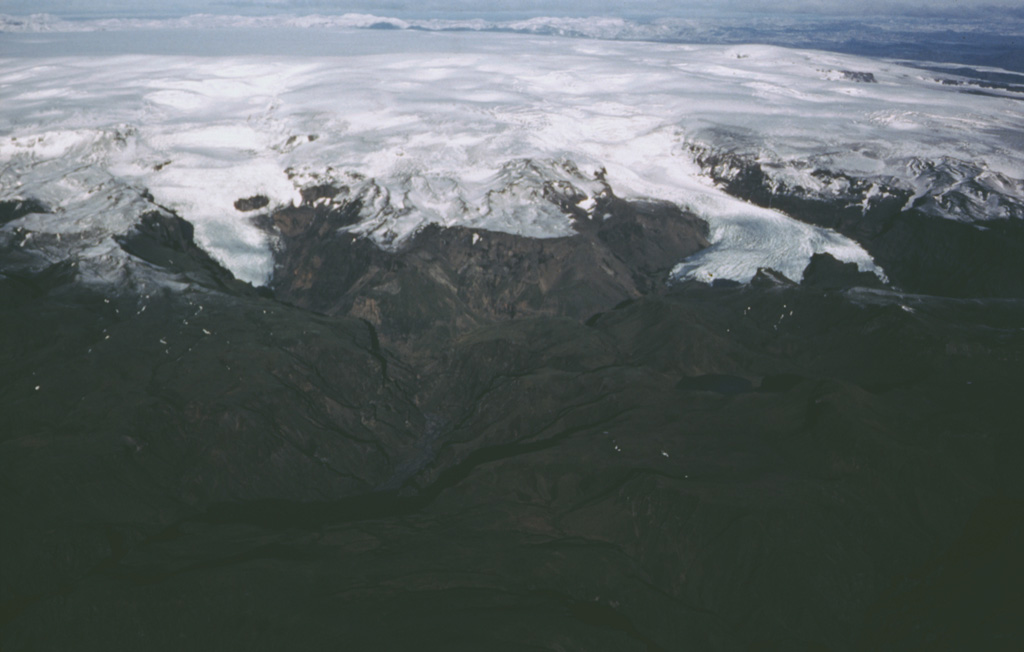 Katla volcano, near the southern end of Iceland's Eastern Volcanic Zone, comprises a central volcano, a 100 km2 caldera beneath the Mýrdalsjökull icecap, plus fissure systems. Valley glaciers descend from the summit toward the coastal plain in this aerial view from the SSW. Katla has very frequent eruptions. Explosive eruptions from the central volcano produce large volumes of tephra and are frequently accompanied by damaging glacier-outburst floods called jökulhlaups. Effusive eruptions producing large volumes of lava are characteristic of the fissure system. Photo by Oddur Sigurdsson, 1985 (Icelandic National Energy Authority).