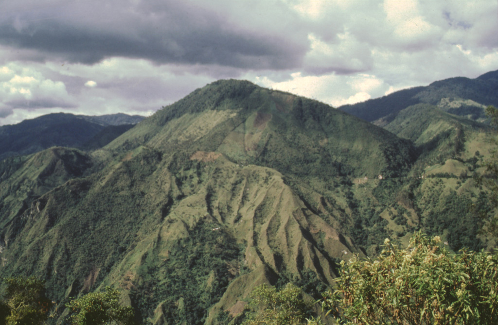 Two central lava domes of Cerro Machín volcano are seen here from the SW. A 3-km-wide caldera, whose rim is the horizontal change in slope across the center of the photo, opens towards the south. Late-Holocene eruptions produced block-and-ash flows that traveled to the west and south, beyond the caldera. Deposits from some of these eruptions form the eroded slopes in the lower part of the photo along the valley of the Toche River. Photo by José Macías, 1996 (Universidad Autómona de México).