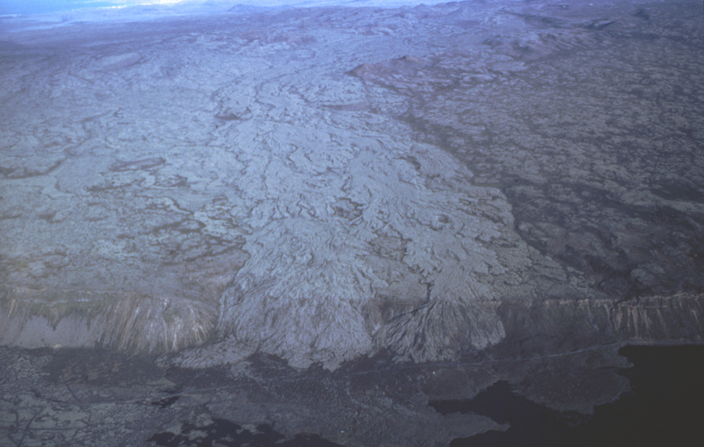 The Brennisteinsfjöll volcanic system, located east of Kleifarvatn lake, consists of a 45-50 km long NE-SW trending fissure swarm, with crater rows and small shield volcanoes. Brennisteinsfjöll is seen here in an aerial view from the south with light-colored lava flows spilling over a 100 m high E-W-trending scarp from vents at least 8 km to the north. The body of water seen bottom left is the Hliðarvatn lake. Photo by Oddur Sigurdsson, 1983 (Icelandic National Energy Authority).