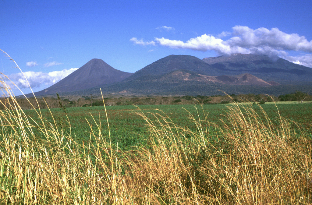 The broad Santa Ana volcano is to the right, with Izalco volcano to the left in this NW-looking view from the Zapotitán basin. The cone in the center is Cerro Verde, and the smaller, low-profile scoria cone below it is Cerro Marcelino. Historical eruptions have occurred from the summit crater of Santa Ana and flank cones such as Cerro Marcelino to the SE. Izalco formed in 1770, and the saddle between Izalco and Cerro Verde increased 100 m in height in the century after 1866. Photo by Lee Siebert, 1999 (Smithsonian Institution).