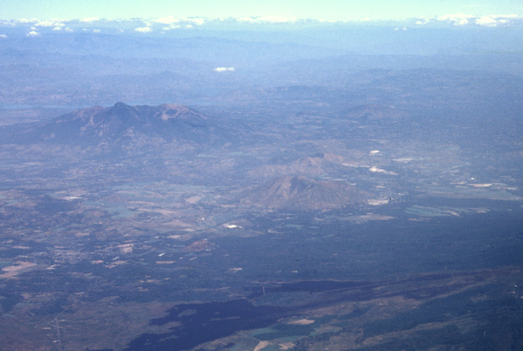 The massive Pleistocene Guazapa stratovolcano (left-center) is seen here in an aerial view from the SW with the Río Lempa behind it.  The youngest flank vent of Guazapa is Cerro Macanze, which lies on the SE flank of the volcano, behind the two small volcanoes in the right-center part of the photo.  The dark-colored unvegetated lava flow in the foreground was erupted in 1917 from the flank of San Salvador volcano. Photo by Paul Kimberly, 1999 (Smithsonian Institution).