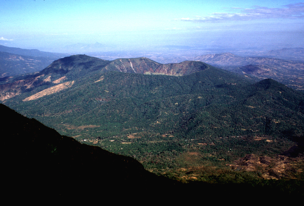 Chinameca stratovolcano is seen here from the SE near the summit of neighboring San Miguel volcano.  A 2-km-wide, steep-sided caldera, Laguna Seca el Pacayal (right-center), truncates the summit of Chinameca volcano.  The Holocene cone of Cerro el Limbo (in the partial shade left of the caldera) on the western flank rises to a point above the level of the caldera rim.  A group of fumarole fields is located on the north flank of the volcano near the town of Chinameca, and the volcano has been the site of a geothermal exploration program.    Photo by Lee Siebert, 1999 (Smithsonian Institution).