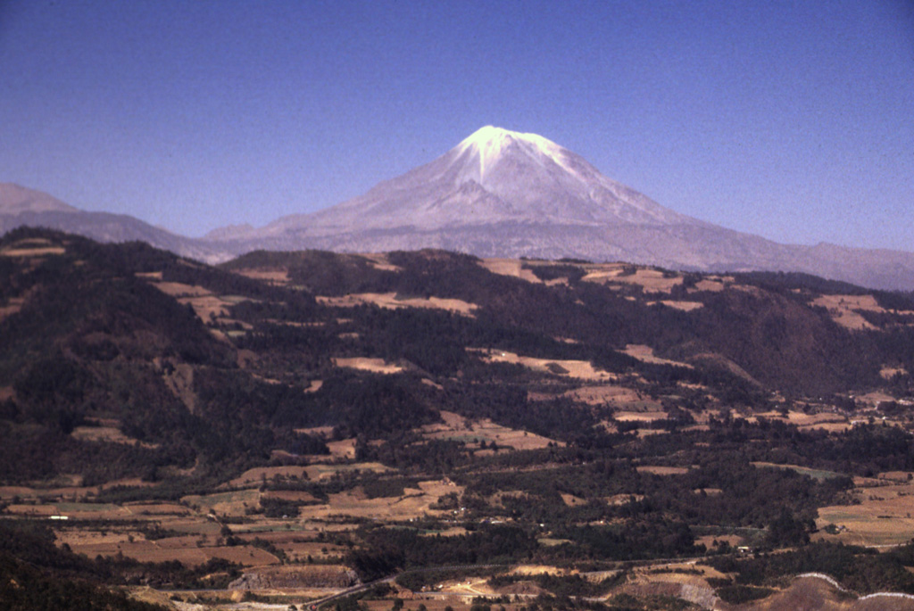 Pico de Orizaba volcano rises above the escarpment at the eastern margin of the Mexican Altiplano. It is seen here from the SE, along the road between Puebla and Orizaba. Like other volcanoes of the Pico de Orizaba-Cofre de Perote chain, deposits of Orizaba are asymmetrically distributed around the summit vent, and extend farther to the east towards the coastal plain.  Photo by Lee Siebert, 1998 (Smithsonian Institution).