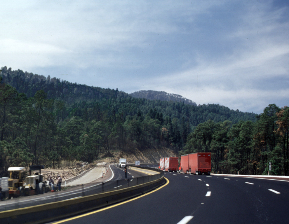 The Mexico City-Puebla highway skirts the northern margin of a voluminous lava flow from Papayo volcano, a lava dome whose rounded summit appears above the horizon to the SW above the trucks to the right. The dome produced massive lava flows that cover an area of 84 km2, with a volume of about 21 km3. The flows display transverse arcuate pressure ridges, and lateral levees. Photo by Lee Siebert, 1999 (Smithsonian Institution).