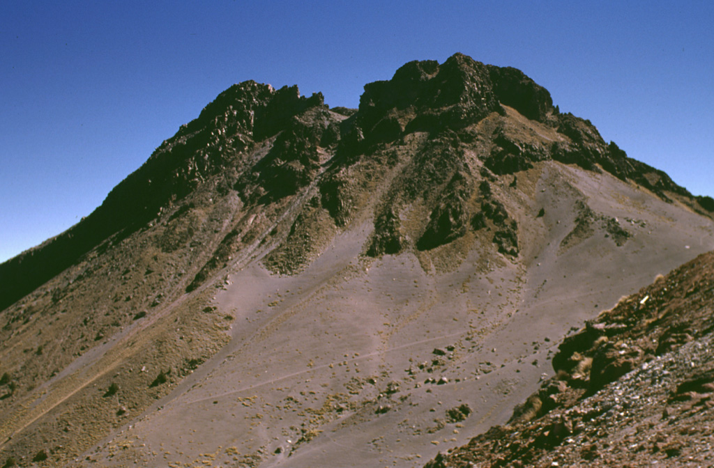 The post-caldera cone of Nevado de Colima forms the high point of the Colima volcanic complex and is the highest peak in western México. The cone, formed of steeply dipping lava flows, was constructed within the youngest of three large scarps that date back to about 500,000 years and has since been extensively glaciated. The scarps open towards the east and formed in part as a result of large flank failures. The youngest is about 5 km in diameter and has a well-preserved western wall, from which this photo was taken. Photo by Lee Siebert, 1998 (Smithsonian Institution).