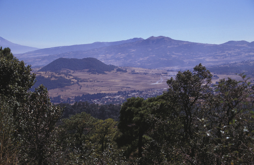 The Chichinautzin volcanic field covers an area of more than 1,000 km2 in a 90-km-wide, E-W-trending area south of the Valley of Mexico. This view looks SE from the 1,600-year-old Xitle scoria cone to Volcán Yololica (the forested scoria cone to left of the center), with Volcán Cuauhtzin on the horizon (right-center) and Volcán Tlaloc to its left. The name Chichinautzin means Burning Lord in the Nahuatl language, and several eruptions have impacted Pre-Hispanic cultures in the Valley of Mexico. Photo by Lee Siebert, 1998 (Smithsonian Institution).