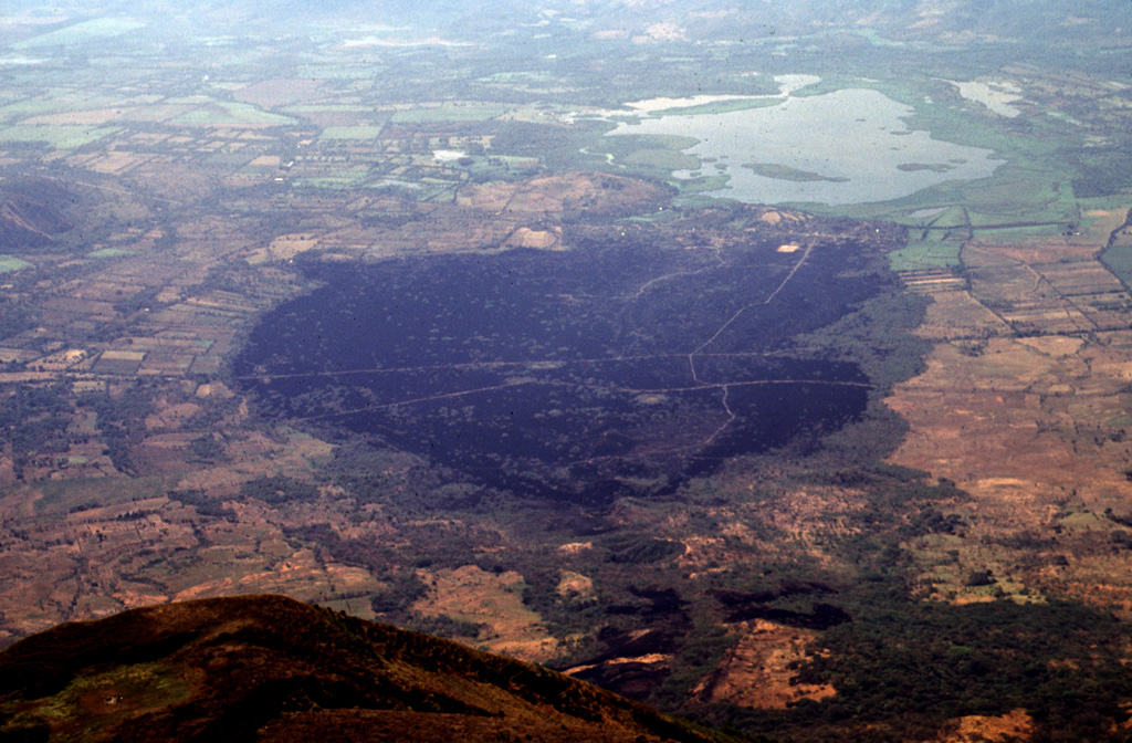 The dark sparsely vegetated area in the center of the photo marks basaltic lava flows erupted on the SE flank of San Miguel volcano in 1819.  The flows, seen here from the summit of San Miguel, originated from fissure vents on the lower SE flank in July 1819 and traveled about 5 km downslope toward Laguna el Jocotal (upper right).  The small lava flows at the lower right-center were erupted in December 1855.  The principal coastal highway and the national railway of El Salvador cross the 1819 flows. Photo by Lee Siebert, 1999 (Smithsonian Institution).