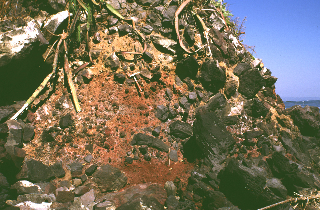 Large clasts in an oxidized scoria matrix of the Acajutla debris avalanche deposit are exposed at the Pacific Ocean coastline more than 40 km from Santa Ana volcano. Clasts with a jigsaw fracture pattern can be seen at the top of the photo and clasts at least 6 m in diameter are nearby. Note the rock hammer in the center for scale. Photo by Lee Siebert, 1999 (Smithsonian Institution).