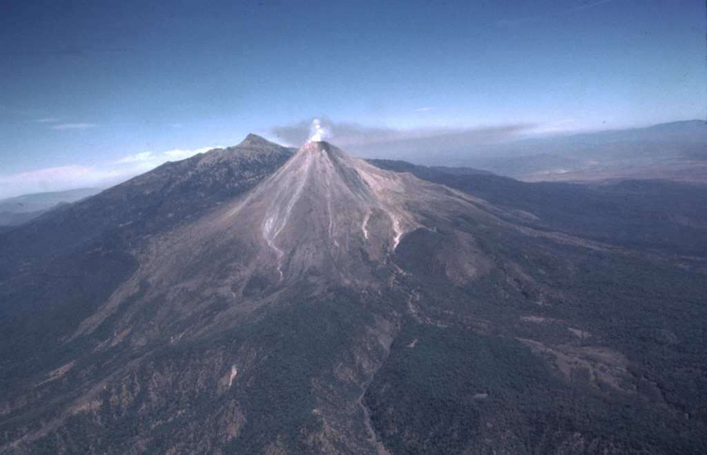 A diffuse gas plume emits from the summit of Colima with a rate of 320 +/- 50 tons/day in February 1982. Volcanism at the Colima volcanic complex has migrated southward from the 300-500 km3 Nevado de Colima in the background. The ancestral Colima volcano was constructed on the southern flank of Nevado de Colima and then collapsed to form the scarp, which now holds the modern Colima edifice (foreground) that grew over the past 2,600 years. Photo by Bill Rose, 1982 (Michigan Technological University).