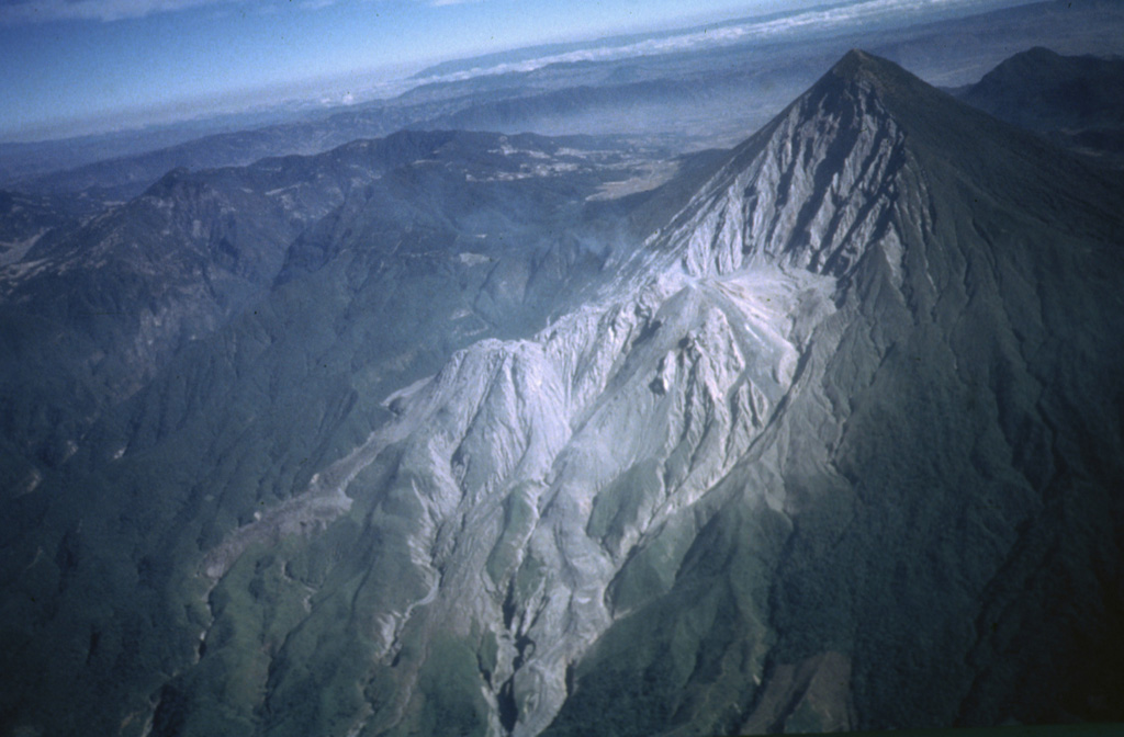 An aerial view from the SE shows the Santiaguito lava dome complex at the base of the 1.5-km-wide 1902 crater on the SW flank of Santa María. Lava flows from the Caliente dome at the eastern side of Santiaguito descend to the bottom center of the photo; lava flows from the western El Brujo vent are visible at the lower left. The broad volcano to the upper left is Siete Orejas.  Photo by Bill Rose, 1980 (Michigan Technological University).