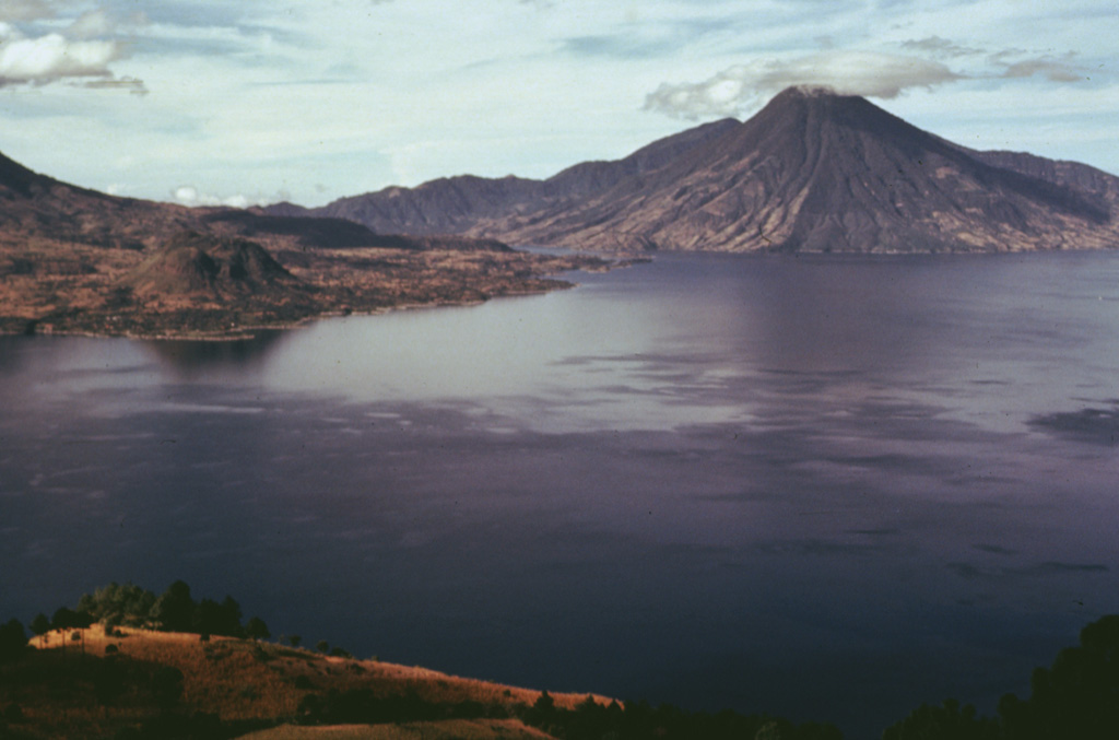 Volcán San Pedro (upper right) is the oldest of three stratovolcanoes constructed within Atitlán caldera. The furrowed slopes of the volcano contrast with the less eroded flanks of the both Atitlán and Tolimán volcanoes. Cerro de Oro, the small lava dome just beyond the shore of Lake Atitlán to the left, is a flank dome of Tolimán. Young lava flows from Tolimán descend into the lake and form part of its southern shore in this view from the west. Photo by Bill Rose, 1978 (Michigan Technological University).