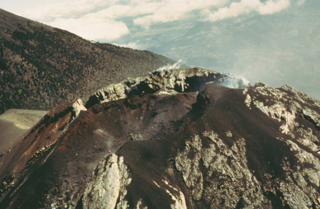 An aerial view of the Fuego summit crater in 1990 shows the products of both effusive and explosive eruptions. The crater walls expose light-colored lava flows and lava agglutinate as well as darker scoria deposits. Historical eruptions have been basaltic in composition, continuing a trend towards more mafic in the Acatenango-Fuego volcanic complex. An Acatenango flank is to the left in this view from the SW. Photo by Bill Rose, 1990 (Michigan Technological University).