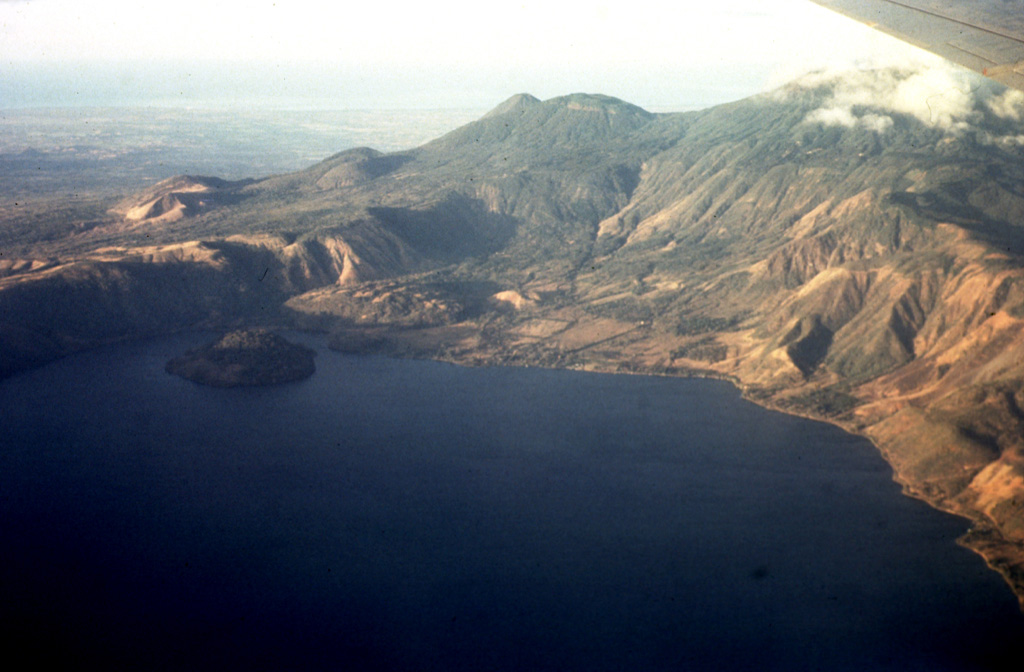 An aerial view of the western side of Coatepeque caldera shows Cerro Grande lava dome, the island to the left. On the ridge behind the caldera wall are (from left to right) the San Marcelino-Cerro la Olla and Cerro el Conejal-Cerro el Astillero complexes, the summit of Izalco volcano, rounded Cerro Verde scoria cone, and (in the clouds) the summit of Santa Ana volcano. The Pacific Ocean is visible in the distance. Photo by Bill Rose, 1967 (Michigan Technological University).