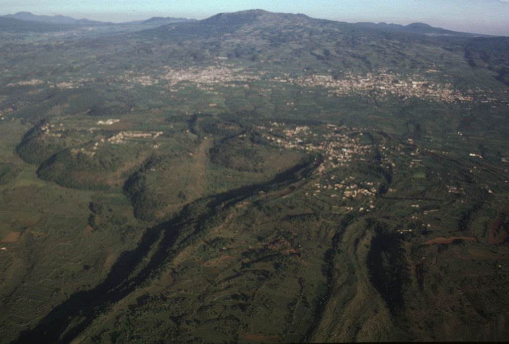 The large, steep-sided Xicomulco lava flow descends from the flanks of the Sierra Chichinautzin into the Valley of Mexico. The viscous lava flow averages 75 m in thickness, displays prominent flow levees, and traveled 4.5 km north. The flow was extruded with very little explosive activity and the vent was subsequently filled in and is now overlain by the town of San Bartolo Xicomulco (right-center). In the background is the town of San Pablo Oztotepec. Volcán Tlaloc is the broad volcano in the center of the horizon. Photo by Gerardo Carrasco-Núñez, 1997 (Universidad Nacional Autónoma de México).