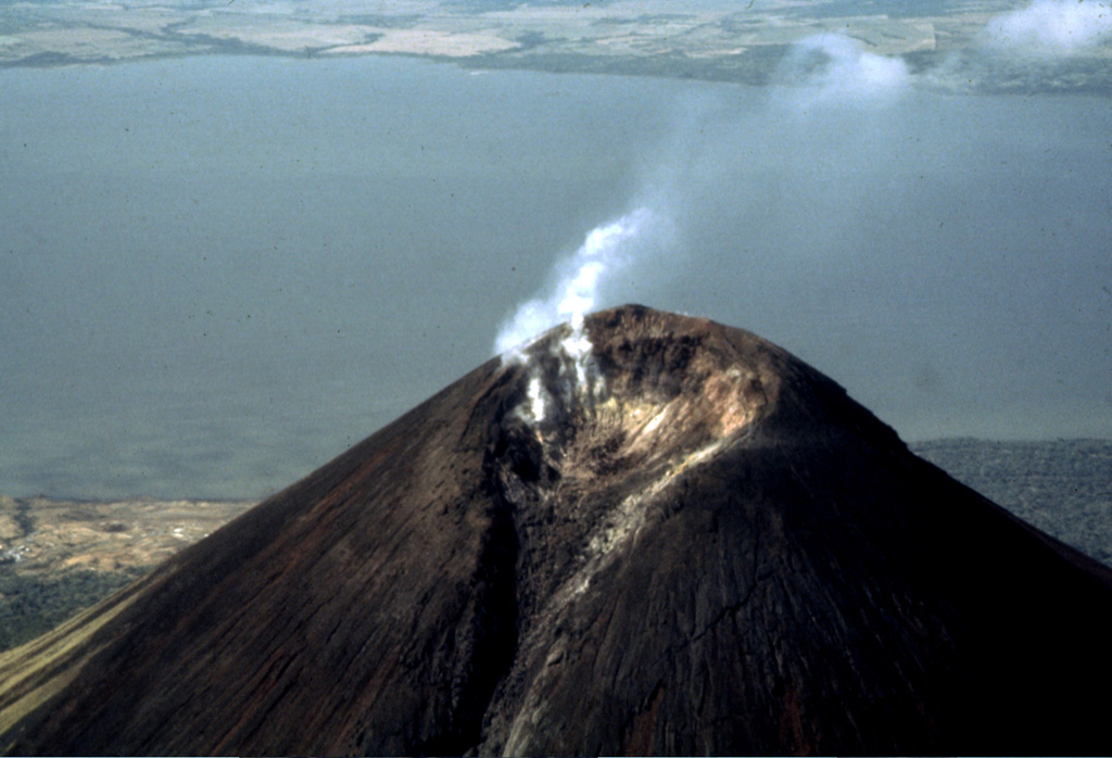 Fumaroles in the summit crater of Momotombo volcano emit vigorous vapor plumes.  The 150 x 250 m wide crater of Momotombo is narrowly breached to the NE, where a lava flow emerged during the 1905 eruption.  The geothermal field on the southern flank of Momotombo can be seen at the lower left along the shores of Lake Managua. Photo by Jaime Incer, 1991.