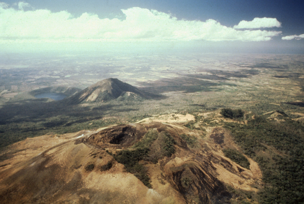 Volcán Las Pilas lies in the foreground in this aerial view of Las Pilas volcanic complex from the north. The diagonal line cutting across from left of the summit crater to the bottom center is the fissure from the 1952 eruption.  The forested crater at the middle right is Cerro Ojo de Agua.  The conical peak in the middle distance is Cerro Asososca, and immediately to its left is Laguna de Asososca maar.  Two other dry maars are located in the Nicaraguan depression between Laguna de Asososca and the tip of Lake Managua at the upper left. Photo by Alain Creusot-Eon, 1980 (courtesy of Jaime Incer).