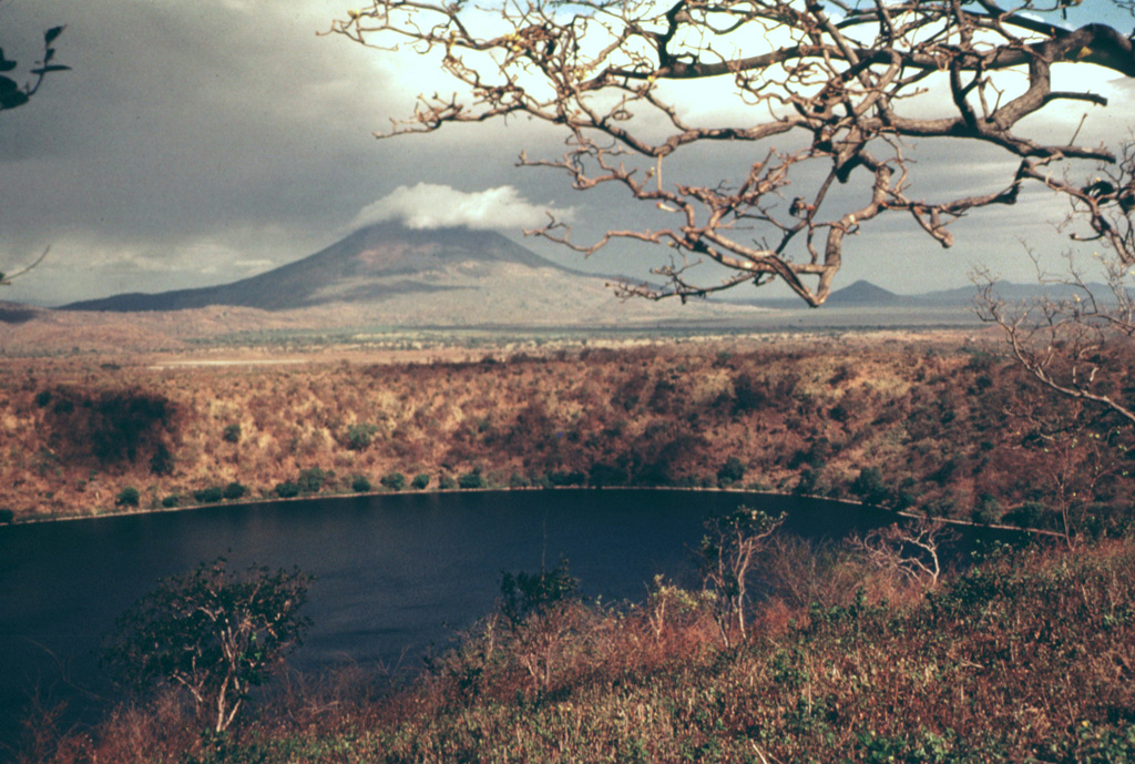 Laguna de Asososca in the foreground is a maar that was constructed along a fissure extending south from Las Pilas volcano.  The maar is elongated N-S in the direction of the fissure system and is about 2 km long in that direction.  Prevailing winds to the west constructed a higher rim on that side, the vantage point of this photo.  The cloud-capped conical peak in the distance is Momotombo, and the much lower peak to its right is Momotombito, which forms an island in Lake Managua. Photo by Jaime Incer.