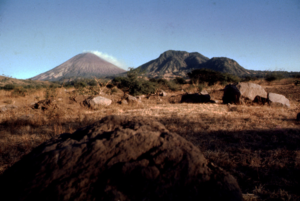 Boulders litter the foreground in this 1977 view of steaming San Cristóbal (left horizon) and Volcán El Chonco (right horizon).  Immediately to the right of El Chonco on its western flank is Loma la Teta, a dacitic lava dome that postdates the collapse.  Another small dome, Loma Caparra (barely visible here left of El Chonco and in front of the steaming San Cristóbal volcano in the background) was extruded on the northern flank of El Chonco.  The 1105-m-high El Chonco lies at the western end of the San Cristóbal volcanic complex. Photo by Alain Creusot-Eon, 1977 (courtesy of Jaime Incer).