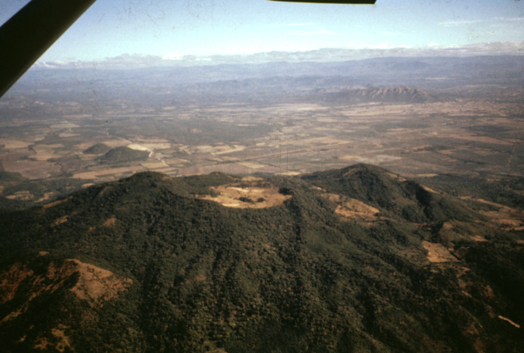 The deeply eroded, forested Rota stratovolcano is seen here in an aerial view from the south.  The vegetated, 832-m-high volcano, also known as Orota, is truncated by a 1-km-wide circular crater.  No historical eruptions are known from Volcán Rota, although seismic swarms occurred in 1986, 1989, and 1992.  Two small NNW-SSE-trending lava domes, El Bosque (also known as Lomas San Ignacio del Bosque or Cerro Ojochal) form the small forested cones visible on the plain at the left center, 2 km north of the flank of Rota. Photo by Jaime Incer, 1991.