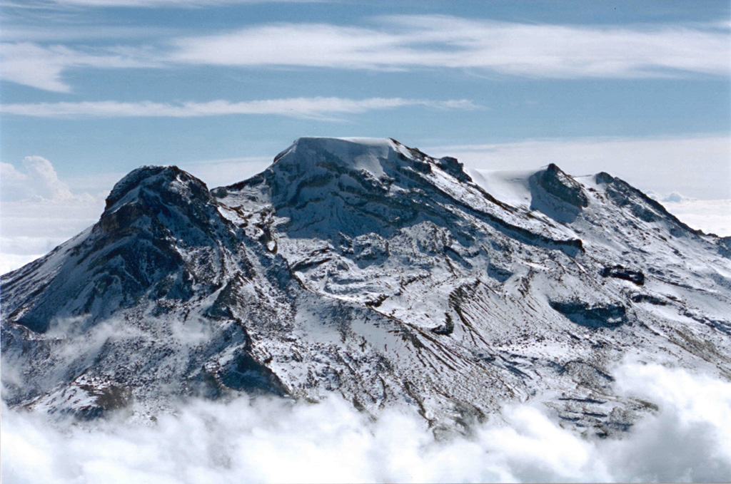 Iztaccíhuatl rises above the clouds in this aerial view from the NW. The summit ridge is formed by a series of overlapping edifices. La Cabeza is to the left, glacier-covered El Pecho in the center, and Las Rodillas to the far right.  Photo by Hugo Delgado, 1996 (Universidad Nacional Autónoma de México).