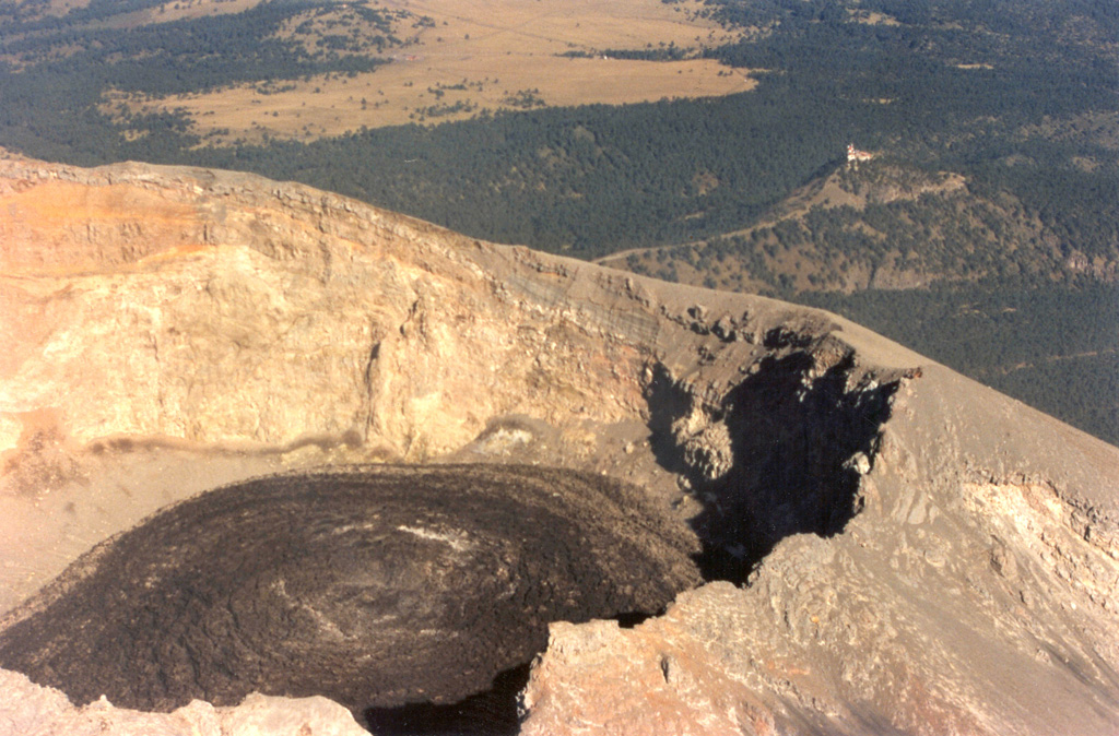 Black lava fills the crater floor of Popocatépetl on 9 December 1997 after rapid lava extrusion on December 5-6 had almost covered the entire crater floor. Circular flow ridges that formed during lava extrusion from the central vent are visible. TV transmission towers at Cerro Tlamacas on the northern flank are seen in the background to the upper right, and at the top of the photo are the light-colored grasslands of Llano Grande at Paso de Cortés, the saddle between Popocatépetl and Iztaccíhuatl volcanoes. Photo by Hugo Delgado-Granados, 1997 (Universidad Nacional Autónoma de México).