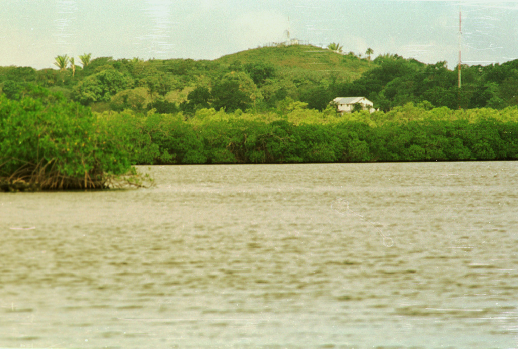 The low peak in the background is Stuert Hill, a pyroclastic cone that rises only 51 m above the Caribbean Sea at the eastern end of Utila Island.  The cone is seen here from across East Harbor at the SE end of the island.  The cone forms an arcuate ridge composed of ejected basaltic tuffs and abundant blocks that include coral fragments, limestone, and metamorphic rocks. Photo by Rick Wunderman, 1999 (Smithsonian Institution).