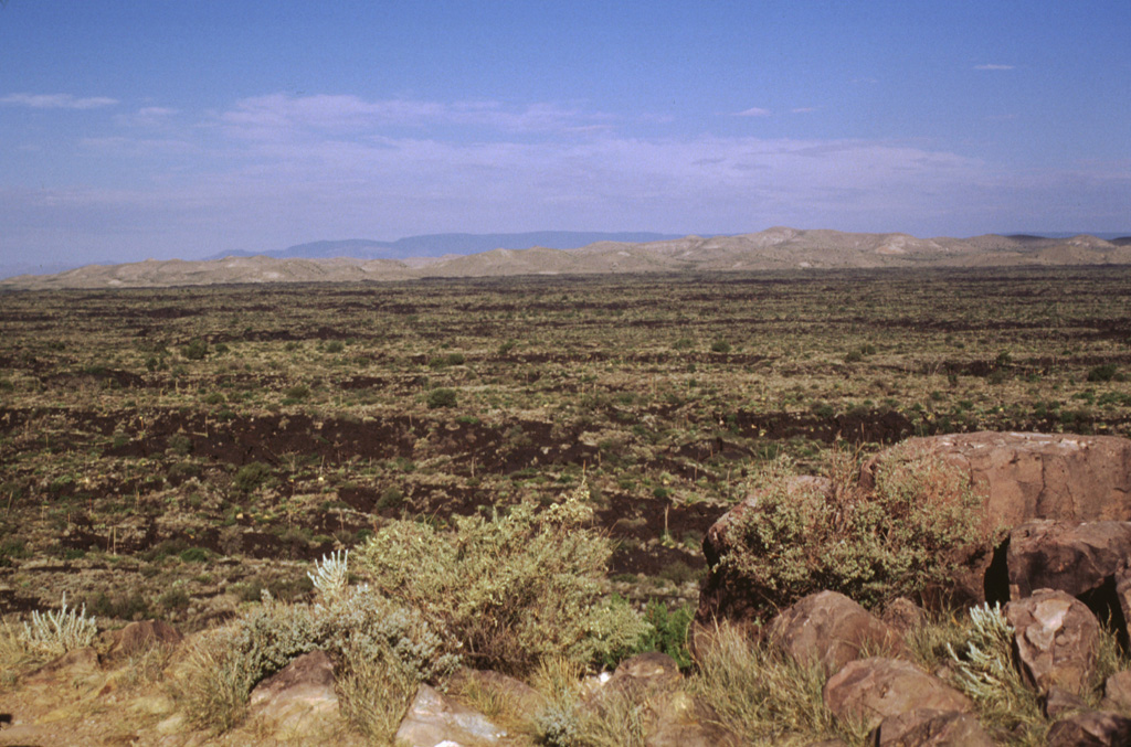 The sparsely vegetated lava flow filling a broad valley is the Carrizozo lava flow, which was erupted from a low shield volcano topped by the Little Black Peak cinder cone.  The massive lava flow, which was dated at about 5200 years Before Present, traveled 75 km down the Tularosa Basin in south-central New Mexico. The extremely lengthy travel distance of the flow (one of the longest on Earth during Holocene time) was facilitated by movement within lava tubes, which thermally insulated the flow.  Photo by Lee Siebert, 1999 (Smithsonian Institution).