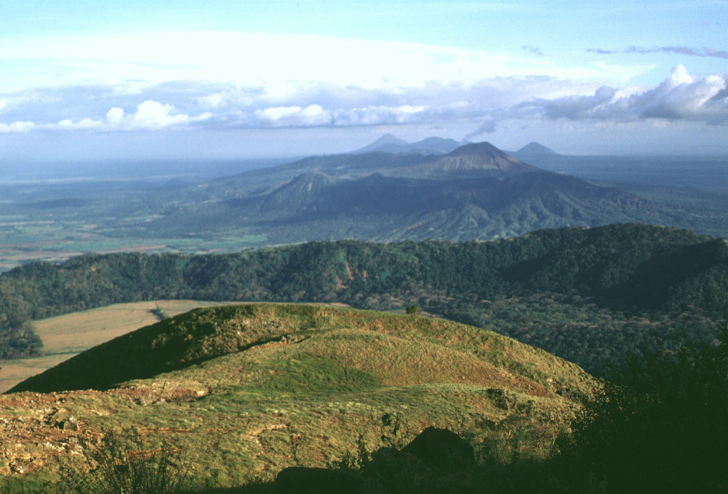 The oldest volcanic center of the San Cristóbal volcanic complex is La Pelona caldera, whose eastern wall forms the forested ridge in the center of the photo.  Formation of the caldera was accompanied by the eruption of large amounts of dacitic pumice.  Part of the caldera floor is visible at the left, but the western rim and floor has been buried by products of Casita volcano, where this photo was taken.  The Marrabios Range volcanoes of Telica, Las Pilas, and conical Momotombo can be seen in the distance. Photo by Lee Siebert, 1998 (Smithsonian Institution).