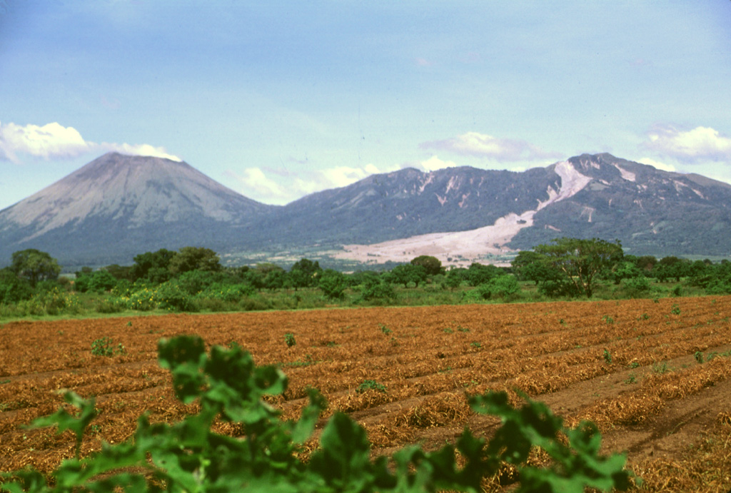 This tranquil scene of steaming San Cristóbal volcano (left) and elongated Casita volcano (right) rising above fields in the León plain is marred by the fresh three-week-old scar of an avalanche and mudflow from Casita.  Torrential rainfall accompanying Hurricane Mitch in October 1998 contributed to the collapse near the summit of Casita, which inundated the towns of El Porvenir and Rolando Rodriguez as well as other settlements. Photo by Lee Siebert, 1998 (Smithsonian Institution).