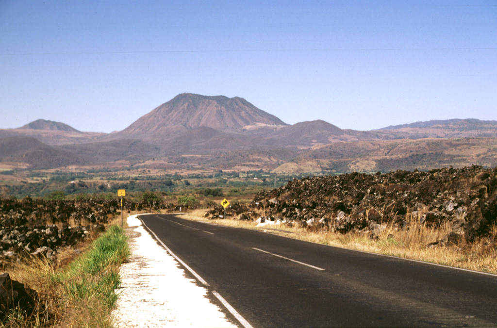 The flat-topped rhyolitic Los Ocotes lava dome (center) was erupted on the southern flank of Tepetiltic volcano about 100,000 years ago and is the youngest product of that complex. Extrusion of the dome blocked drainages to form Laguna San Pedro, out of view to the left. The rim of Tepetiltic caldera forms the right horizon. Los Ocotes is seen here from the SE along Highway 15 where it crosses the blocky andesitic El Ceboruco lava flow in the foreground, which traveled 8 km from Ceboruco volcano. Photo by Lee Siebert, 1997 (Smithsonian Institution).