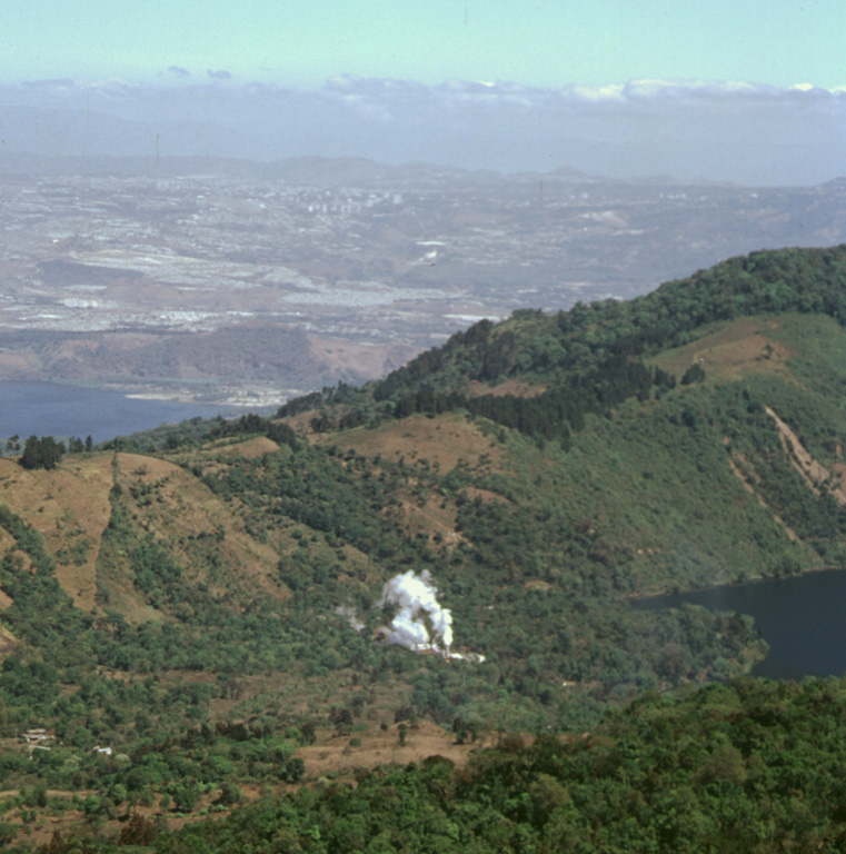A steam plume rising from a geothermal site in Amatitlán caldera is seen here from the caldera rim of Pacaya, with Guatemala City in the background. Laguna Calderas contains the lake to the right and beyond the ridge to the left is part of Lake Amatitlán. The 14 x 16 km Amatitlán caldera produced a large number of major explosive eruptions that covered the current site of Guatemala City with pyroclastic flows during the late Pleistocene. The caldera retains a high heat flow that is being exploited for geothermal energy. Photo by Paul Kimberly, 1999 (Smithsonian Institution).