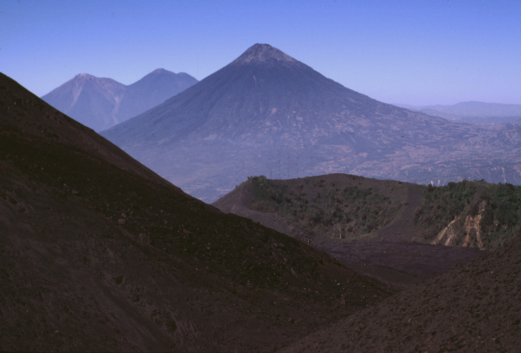 Volcán de Agua is in the center of this photo and lava flows from MacKenney cone form the slope in the foreground. They have filled in the moat of the Pacaya caldera almost to the level of the lower Cerro Chino crater rim, with the communication antennas along the summit in the midground. The two volcanoes on the left horizon are Fuego (left), one of the most active in Guatemala, and Acatenango (right). Photo by Lee Siebert, 1999 (Smithsonian Institution).