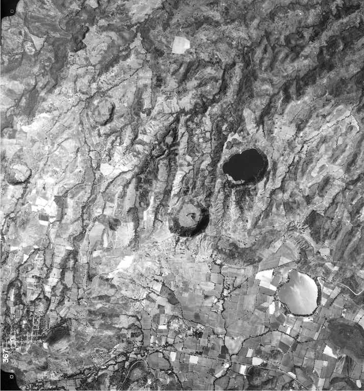 Several maars of the Apastepeque volcanic field are seen in this vertical aerial view.  The two lake-filled maars are Laguna de Apastepeque (the light-colored lake at the lower right) and Laguna Chalchuapa (the dark-colored lake at the right-center).  SW of it is the large dry maar Hoyo de Calderas, and other dry maars appear at the upper left and lower right.  The Pan-American highway cuts across the bottom of the photo, and the town of San Esteban Catarina is at the lower left, immediately west of Cerro Las Delicias lava dome.  Aerial photo by Instituto Geográfico Nacional El Salvador, 1992.