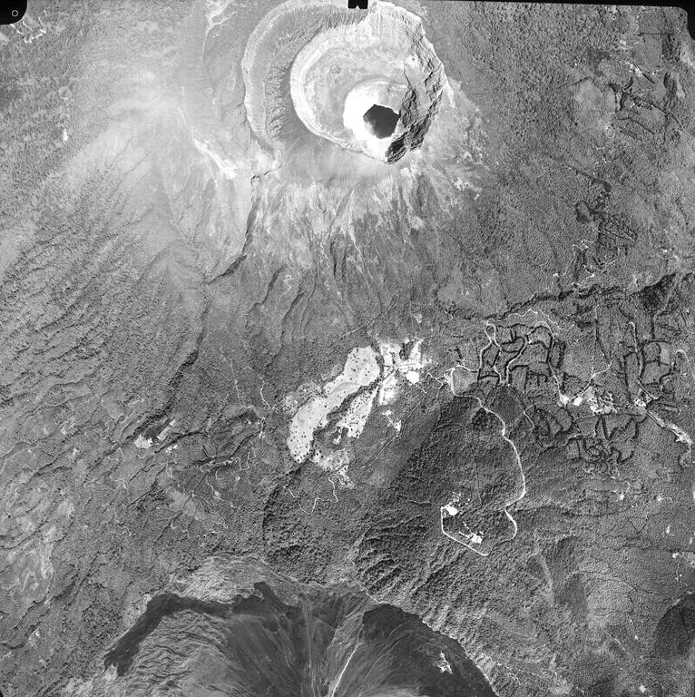 A lake fills the youngest of four nested craters of Santa Ana volcano (top center).  A road winds at the lower right to radio antennas at the summit of forested Cerro Verde cinder cone, one of a series of volcanic cones constructed along a NNW-SSE-trending fissure cutting across Santa Ana.  The dark unvegetated areas at the bottom of the photo are the northern flanks of Izalco volcano. Aerial photo by Instituto Geográfico Nacional El Salvador, 1996.