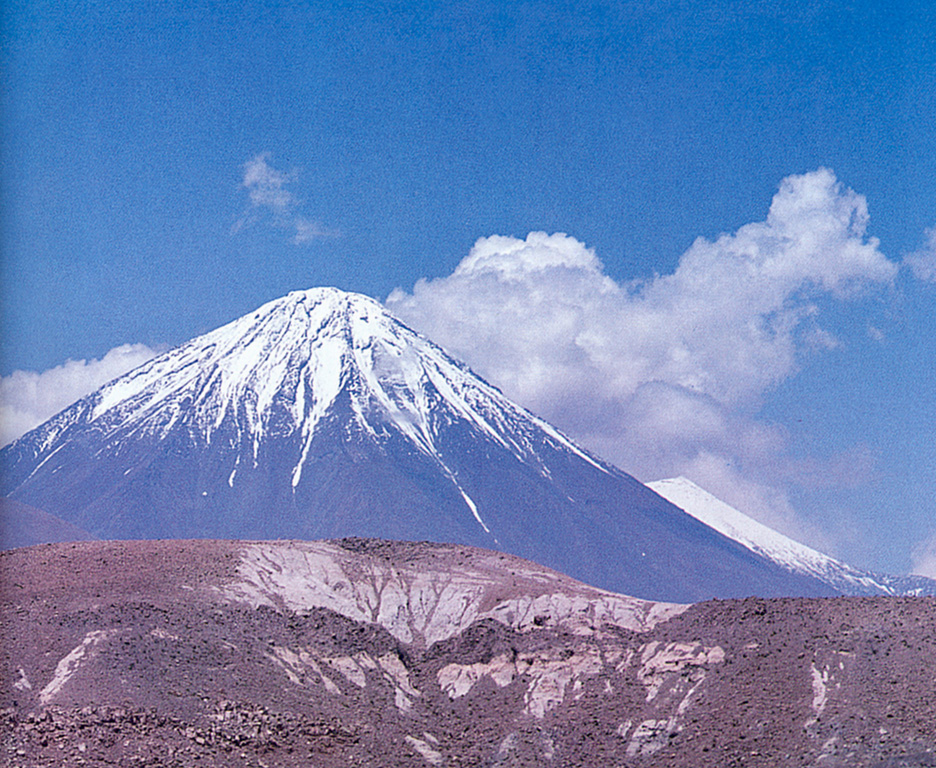 Snow-capped Licancabur volcano rises to the east beyond a Pliocene rhyolitic pyroclastic-flow deposit in the foreground from the Chaxas lava dome.  Block lava flows from Licancabur have traveled as far as 12 km from the summit crater. Photo courtesy of Oscar González-Ferrán (University of Chile).