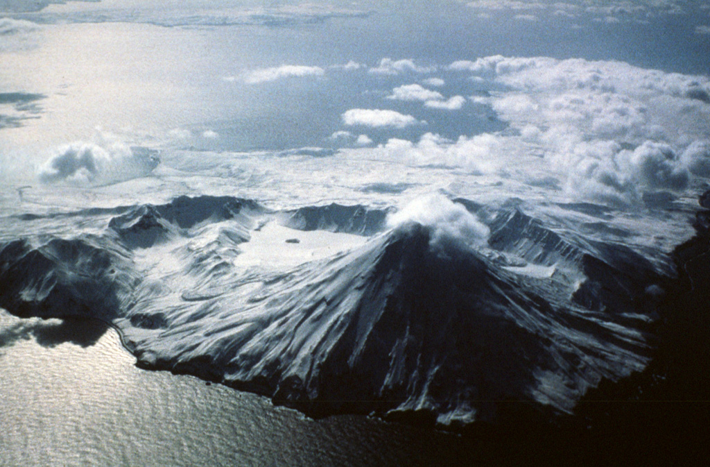 The arcuate ridge in the center of the photo is the southern rim of Kanaton caldera that contains a snow-covered lake in the SE part (center) with a small island. The post-caldera Kanaga cone is in the foreground. Recent lava flows descend the flanks, particularly on the SW, S, and E. Photo by Fred Deines, 1993 (U.S. Fish & Wildlife Service, courtesy of Alaska Volcano Observatory).