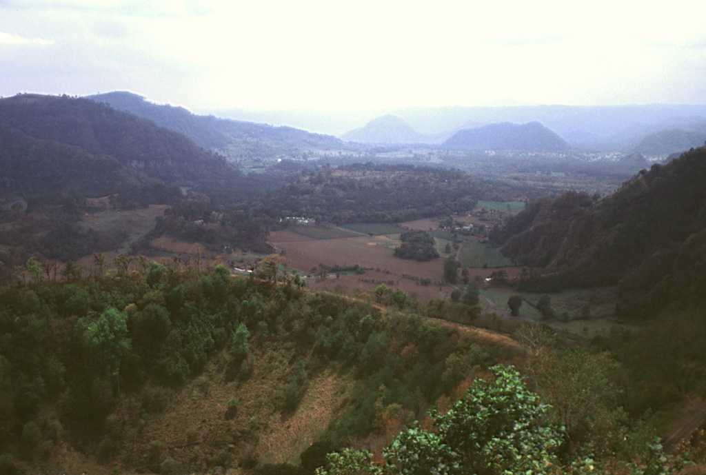 The Coacoatzintla lava flow that fills this valley floor originated from a vent at the base of Rincón de Chapultepec crater in the foreground. It was dated to about 3,000 years ago and underlies the present-day town of Coacoatzintla, seen in front of the dark-colored hill below the right horizon. The flow, which traveled 6 km to the south, diverted around the hill (formed of limestone bedrock) into the valley of the Río Naolinco, which extends from right to left in the distance. This eruption is the youngest known from the Naolinco Volcanic Field. Photo by Lee Siebert, 2000 (Smithsonian Institution).