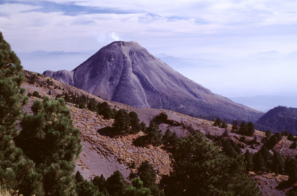 A weak gas plume rises from Volcán de Colima, seen here from the north near the antenna station on Nevado de Colima. The prominent lava flow descending the center of the north flank was erupted in 1961 at the time of the initial overflow of the 1913 crater. The ridge on the left-hand slope is El Volcancito, a NE-flank lava dome that began growing in 1869. Photo by Lee Siebert, 2000 (Smithsonian Institution).
