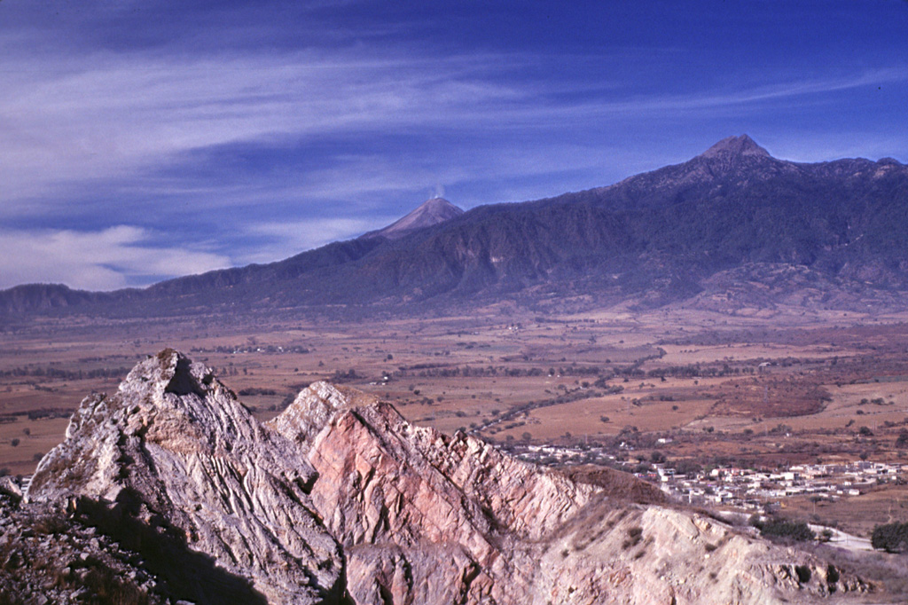 Volcán de Colima (center) and Nevado de Colima (right) are seen here from a quarry at Cerro la Escalera, 23 km NE. Up to 25 m of volcanic deposits (not visible in this photo) from both Cántaro and Colima volcanoes are exposed at Cerro la Escalera. The rocky outcrop in the foreground is a quarry wall cut in Cretaceous limestones for cement. The town of Huescalapa is visible to the lower right within the Colima graben. Photo by Lee Siebert, 2000 (Smithsonian Institution).