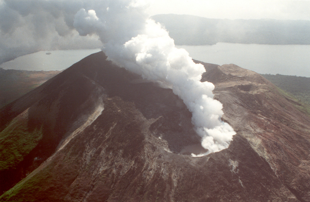 The summit crater complex of Mt. Garat with Lake Letas in the background, seen here from the S. A plume rises from the SE crater. Strong continuous degassing began following an increase in fumarolic activity in April 1991 and was continuing at the time of this July 1991 photo. Mt. Garat is located within the 6 x 9 km wide caldera that is partially filled by Lake Letas. The northern caldera rim is in the background. Photo by Claude Robin, 1991 (ORSTROM, Vanuatu).