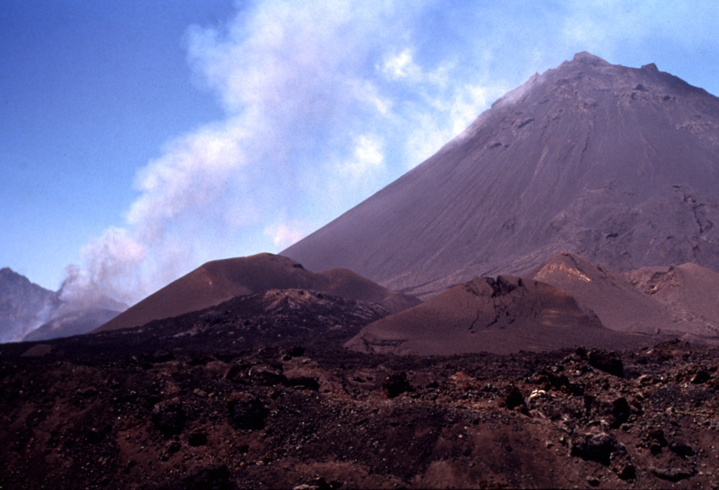 A steep-sided central cone, Pico do Fogo, rises more than 1 km above the floor of the Cha caldera. The island of Fogo is a massive stratovolcano, truncated by a 9-km-wide caldera that is breached to the east. Pico was apparently in almost continuous activity from the time of Portuguese settlement in 1500 CE until around 1760 CE. It is seen here in 1995 during an explosive and effusive eruption from a vent on its lower western flank (left). A plume of ash and steam is visible from the vent. Fogo erupted again in 2014-2015, when lava flows destroyed the intra-caldera villages of Portela and Bangaeira. Photo by Nicolau Wallenstein, 1995 (Center of Volcanology, Azores University).