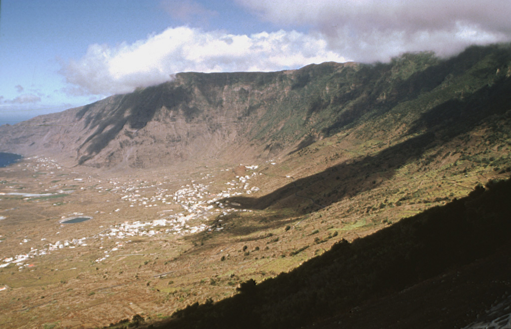 The massive El Hierro shield volcano is truncated by a large NW-facing escarpment, seen here, which formed as a result of gravitational collapse of the volcano. The steep-sided 1,500-m-high scarp towers above a low lava platform bordering 14-km-wide El Golfo Bay, which is barely visible at the extreme left. Holocene cones and flows are found both on the outer flanks and in the El Golfo depression. The latest subaerial eruption, during the 18th century, produced a lava flow from a cinder cone on the northwest side of El Golfo. A submarine eruption occurred in 2011-12 near the southern flank. Photo by Yasuo Miyabuchi, 1997 (Forestry and Forest Products Research Institute, Kyushu).