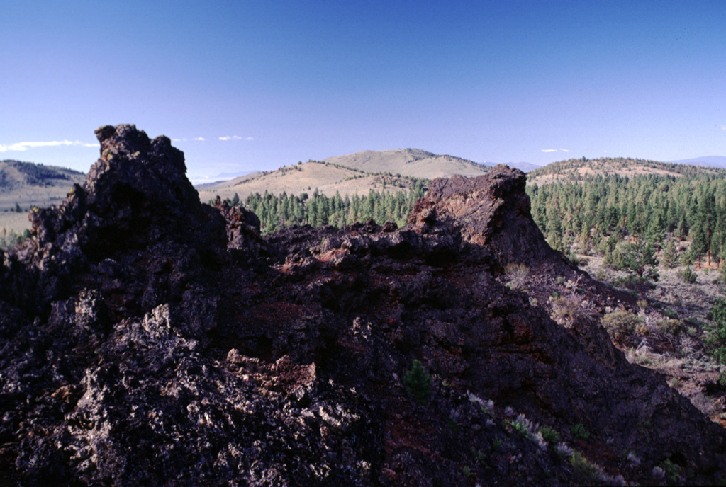 The Blowouts, the spatter vents in the foreground, are the source vents of the voluminous Devils Garden lava field. Devils Garden is the NW-most of a group of three lava fields SE of Newberry volcano and contains 117 km2 of overlapping pahoehoe lava flows erupted from fissure vents at the NE part of the field. The extremely fluid and inflated pahoehoe lavas typically left flows that increase from about a half meter thickness near the vent to about 5 m in the distal portion. The lava flows are either late Pleistocene or early Holocene in age. Photo by Lee Siebert, 2000 (Smithsonian Institution).