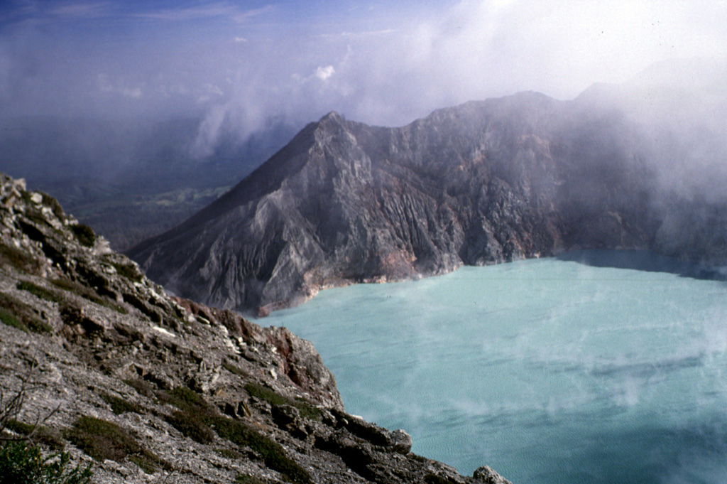 The steaming surface of Kawah Ijen crater lake from the southern rim in July 2000. The outlet of the lake is blocked by a small dam to the left. The maximum depth of the lake in 1996 was 180 m and the volume of the lake had gradually decreased from 0.044 km3 in 1922 to 0.036 km3 in 1996. The location of the most active vents below the lake surface have shifted to the W, towards the dam. Photo by Lee Siebert, 2000 (Smithsonian Institution).