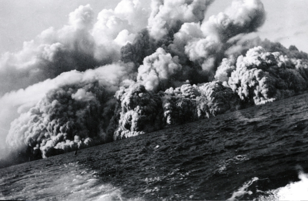 Pyroclastic surges at about 0805 on 1 August 1952 travel across the sea at the western coast of the island, 20 minutes after the start of the eruption. Several inches of ash and lapilli up to 12 mm in diameter fell on the deck of the fishing boat from which this photo was taken as it sailed away from the volcano at full speed. Photo by Robert Petrie, 1952 (U.S. Navy; courtesy of Sherman Neuschel, U.S. Geological Survey).